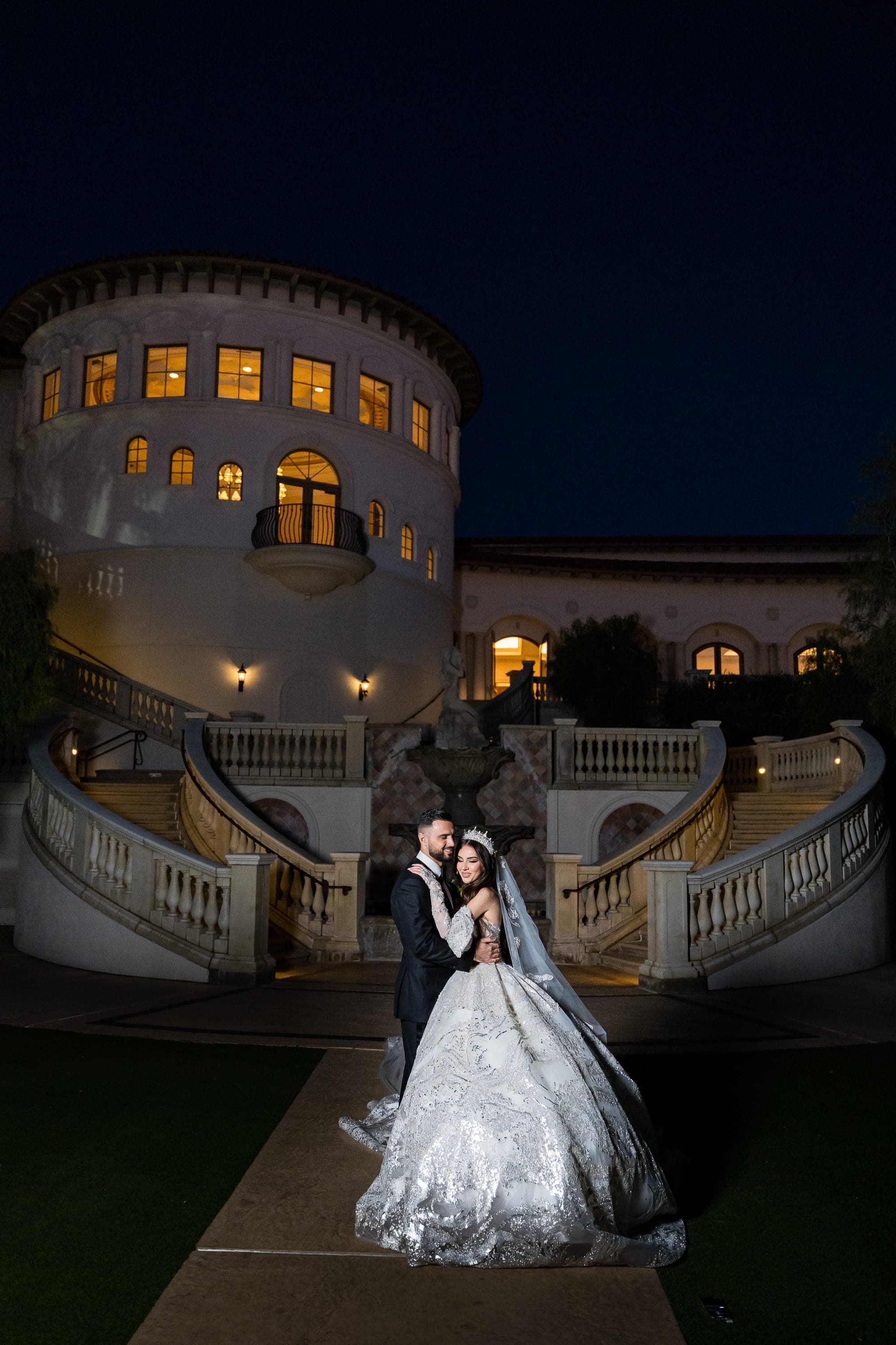 Bride in a sparkling silver gown embraces her groom before a lit mansion with curved staircases at night.