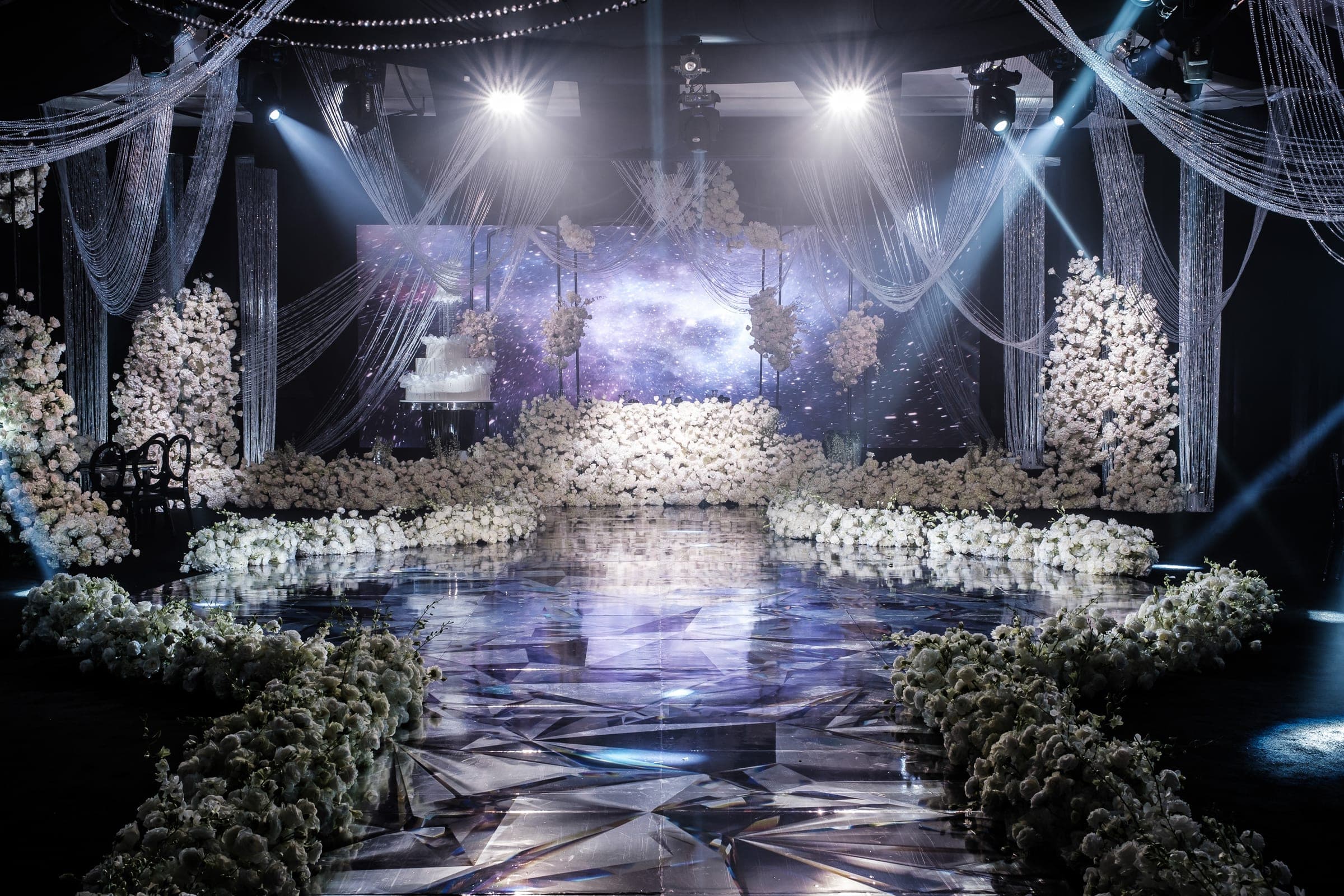 Elegant reception stage flanked by massive white flower walls and a suspended cake under galaxy screen lighting.
