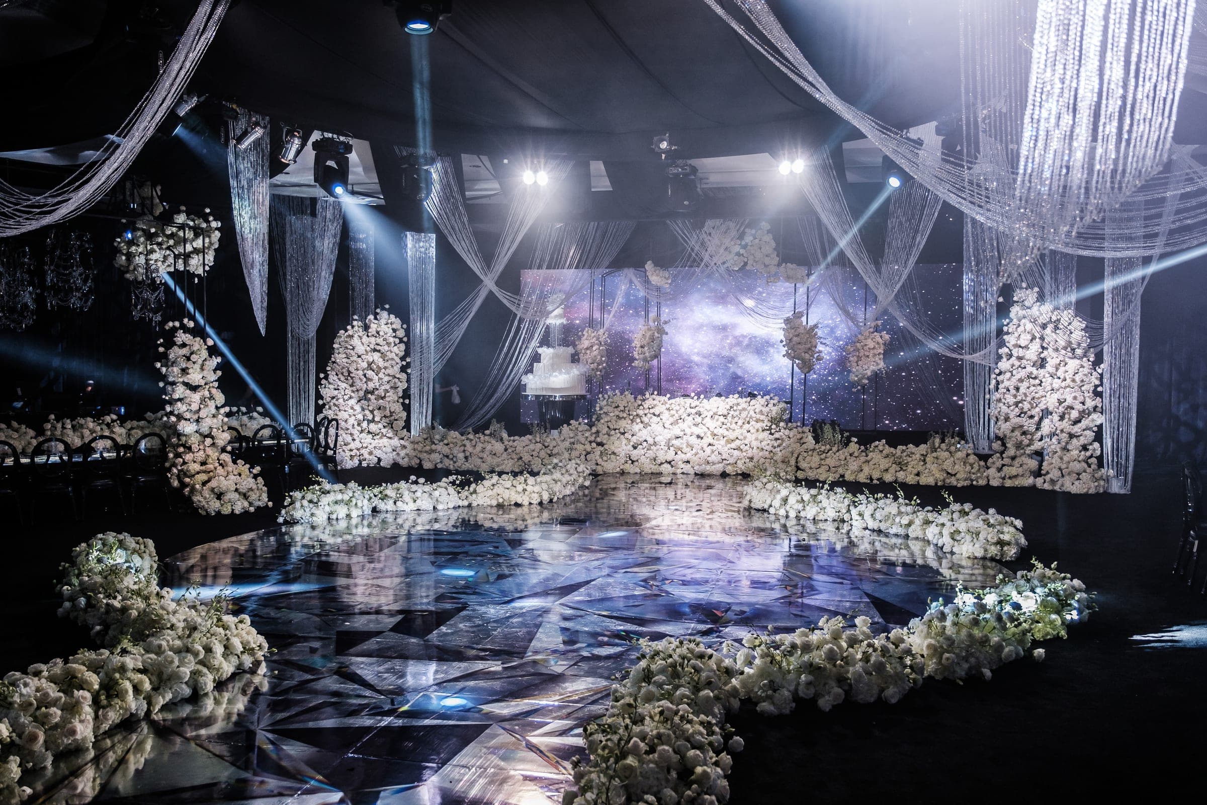 Wide reception hall with geometric mirrored floor, white rose borders, and dangling crystal drapery under spotlights.