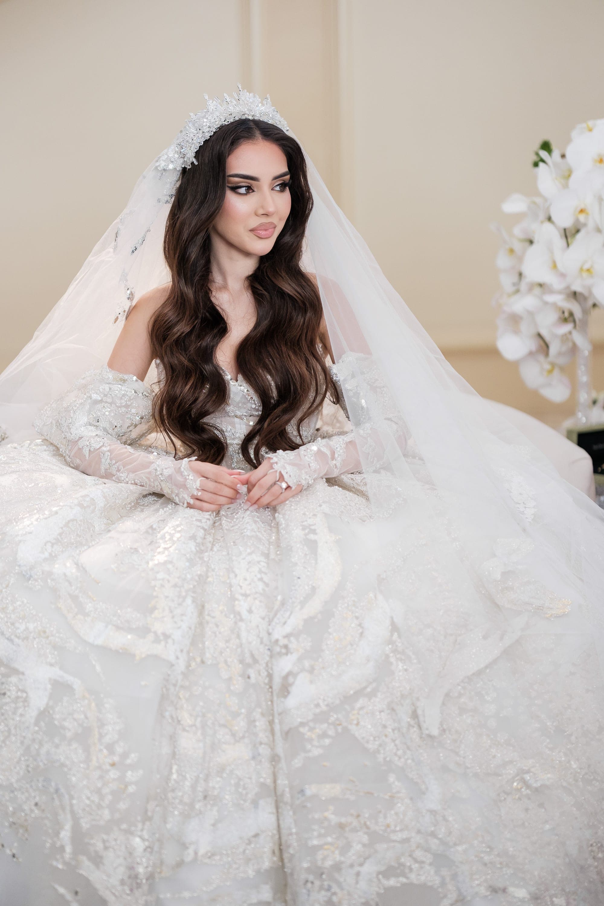 Bride in a sparkling ball gown and crystal crown sits with long dark waves and a flowing veil.