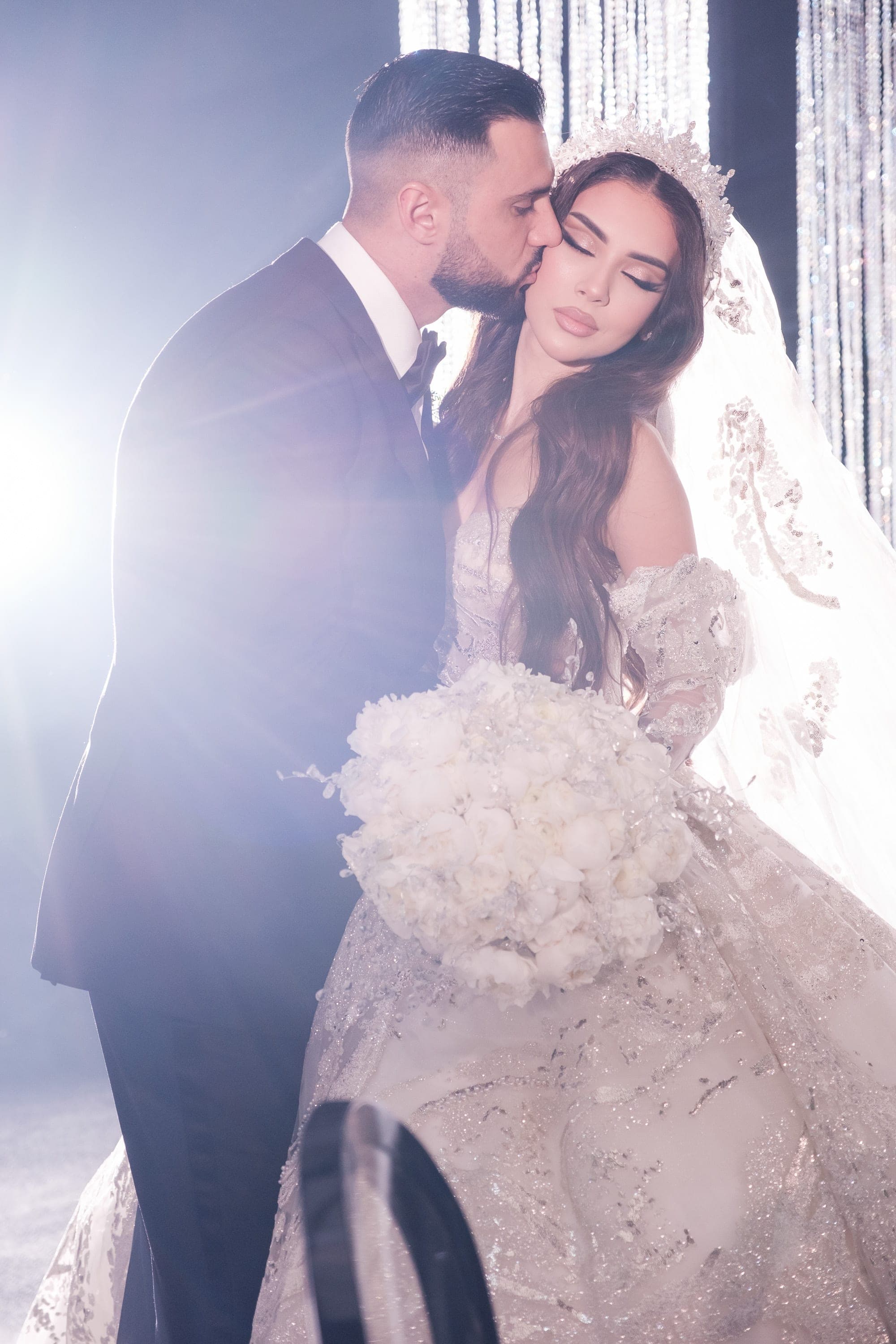 Groom in a black tuxedo kisses the bride's cheek as bright backlight glows behind them.