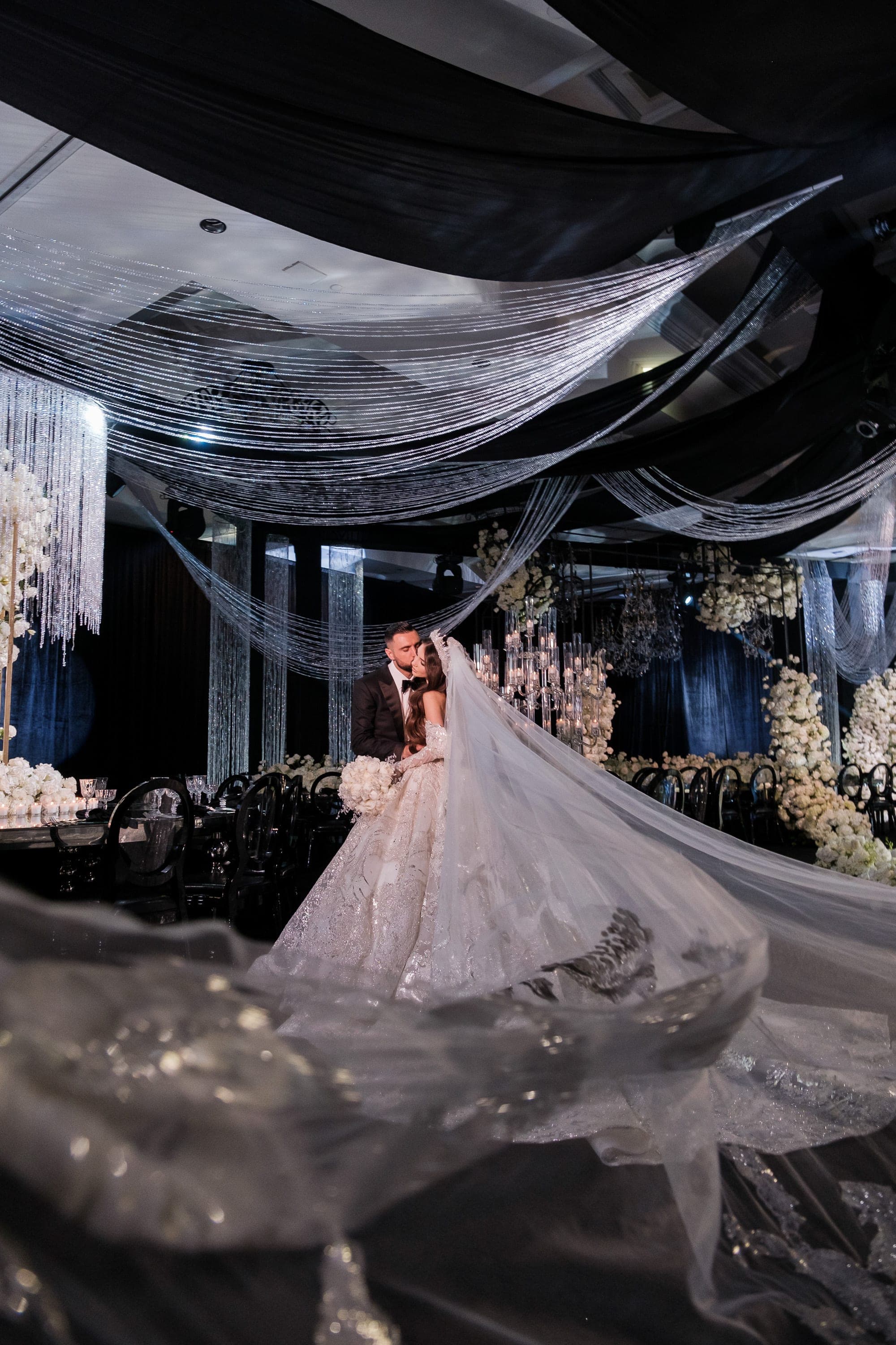 Couple embraces beneath draped fabric and beaded chandeliers in a dark elegant ballroom with white florals.