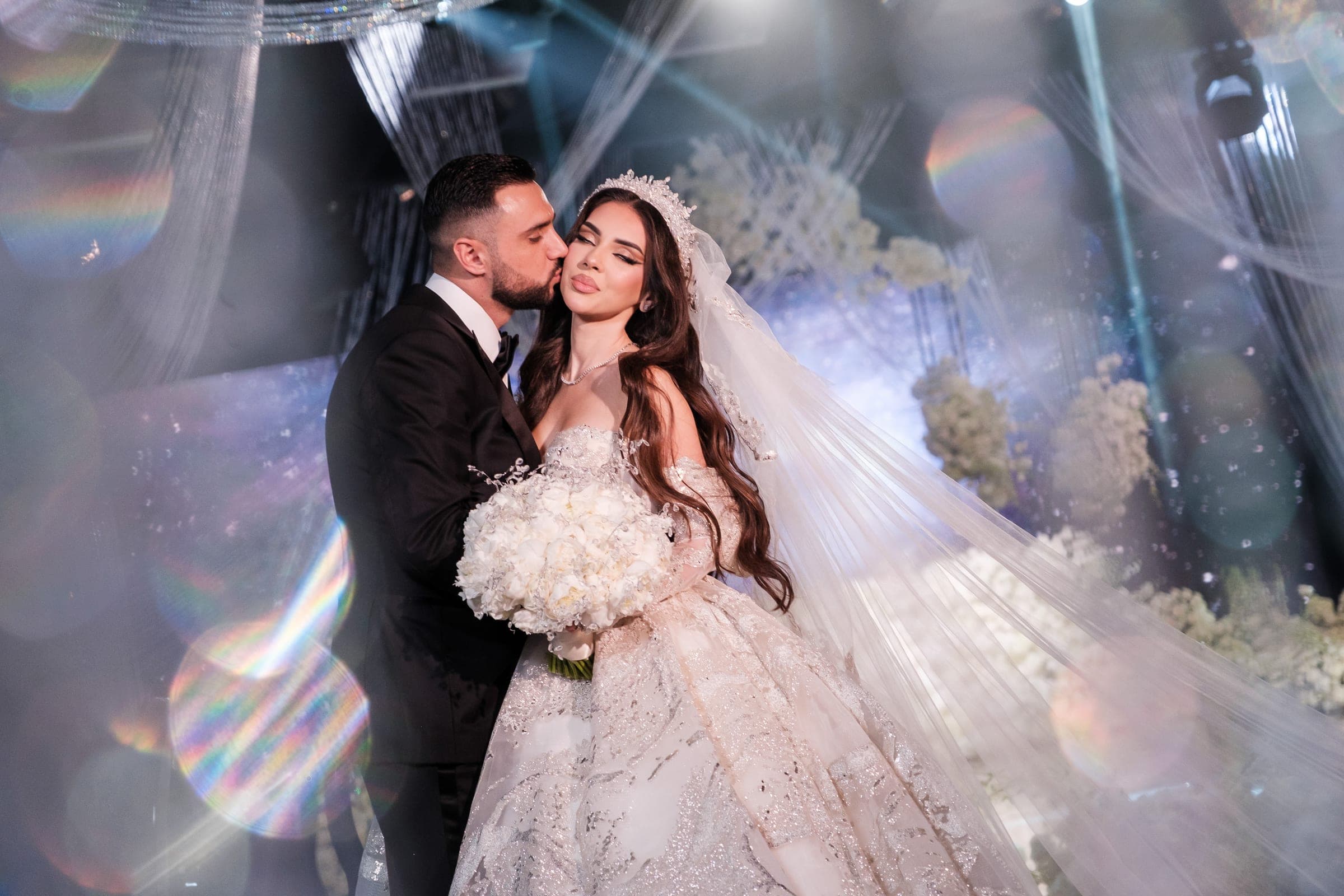 Groom kisses the bride's temple as she holds a white bouquet amid colorful light flares.