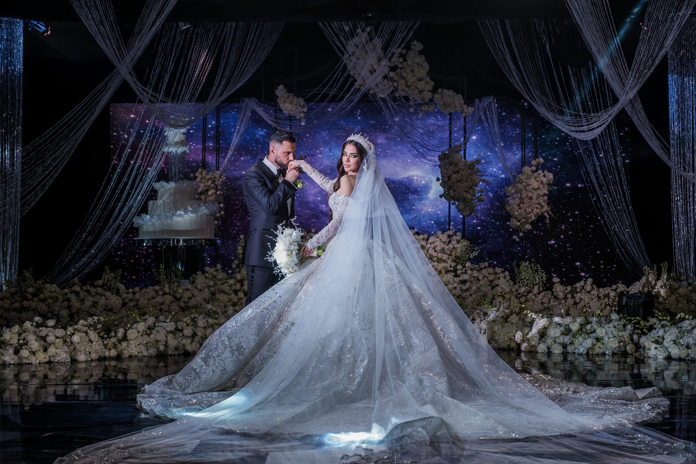 Groom kisses the bride's hand before a galaxy backdrop framed by white flowers and cascading strands.