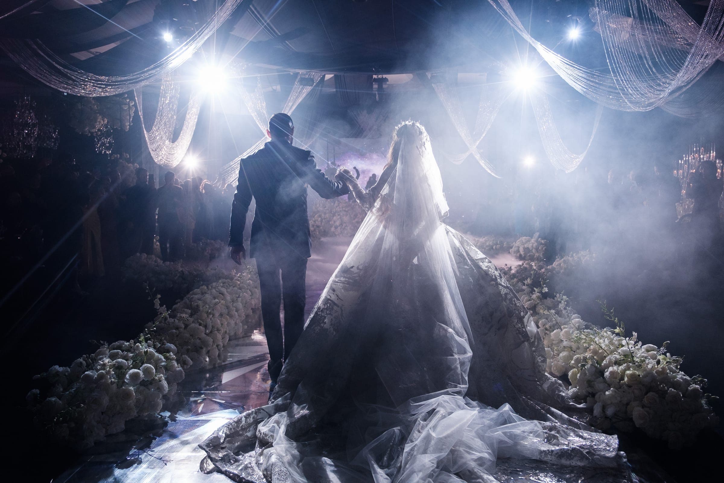 Silhouetted couple walks hand in hand down a misty flower-lined aisle under bright spotlights.