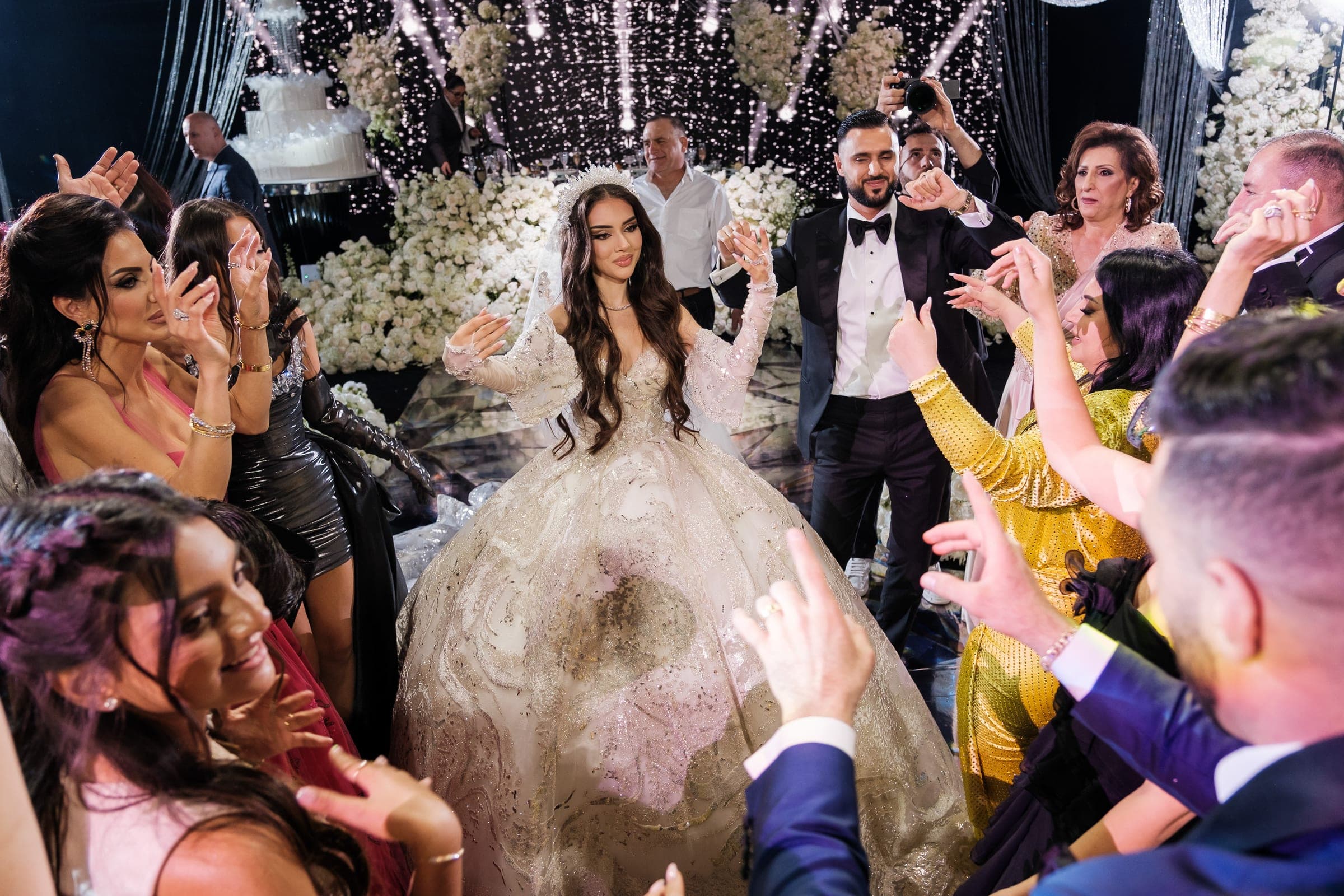 Bride dances joyfully surrounded by clapping guests with a tall cake and white florals behind.