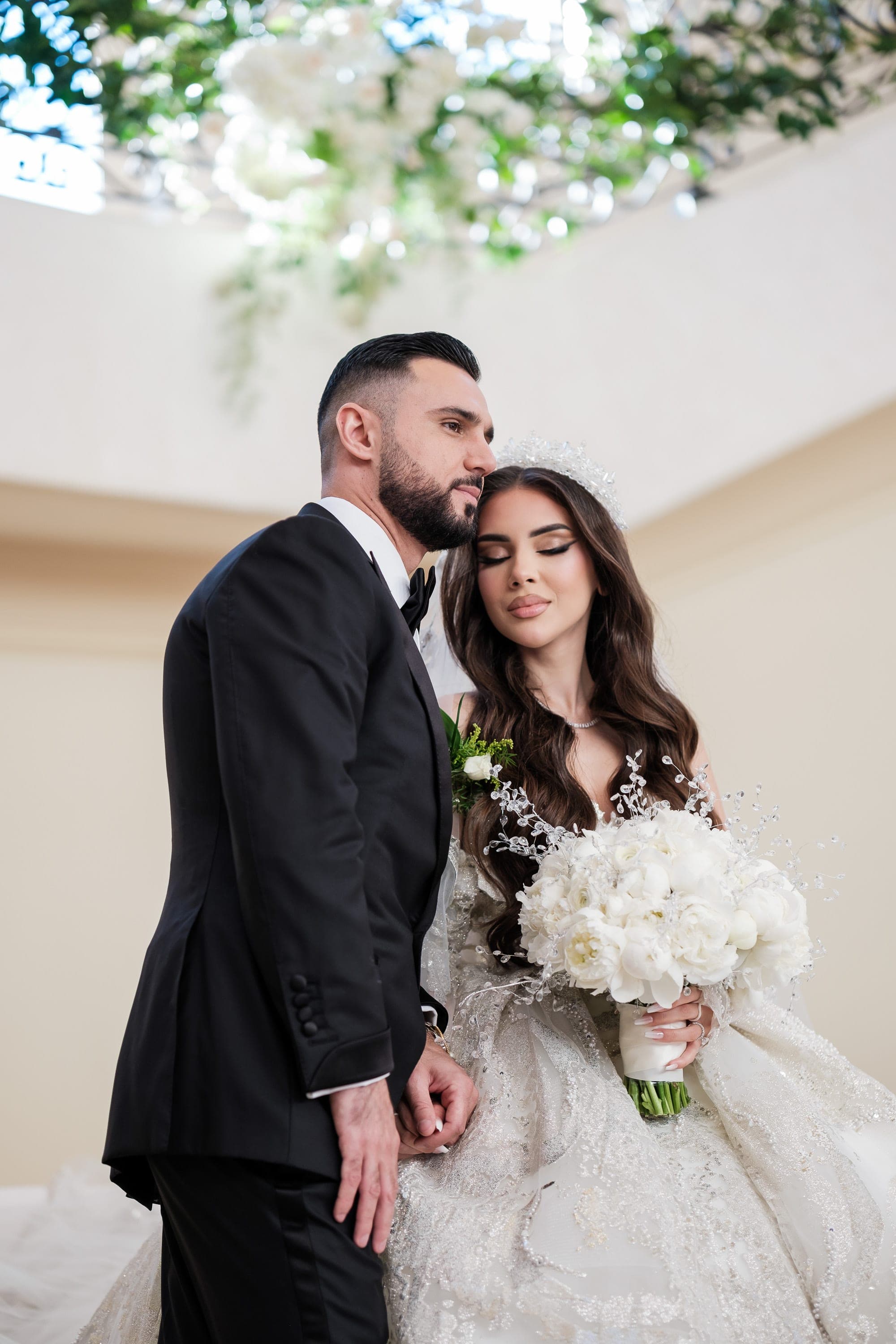 Groom in black tuxedo leans close to bride holding a lush white bouquet beneath hanging blooms.