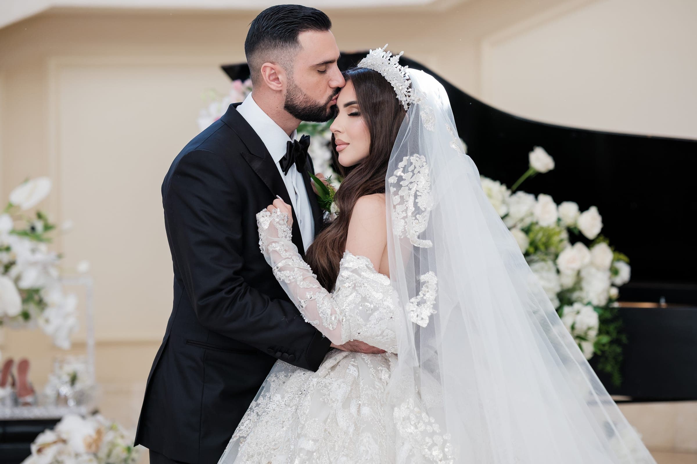 Groom kisses bride's forehead as she closes her eyes near a grand piano and white florals.