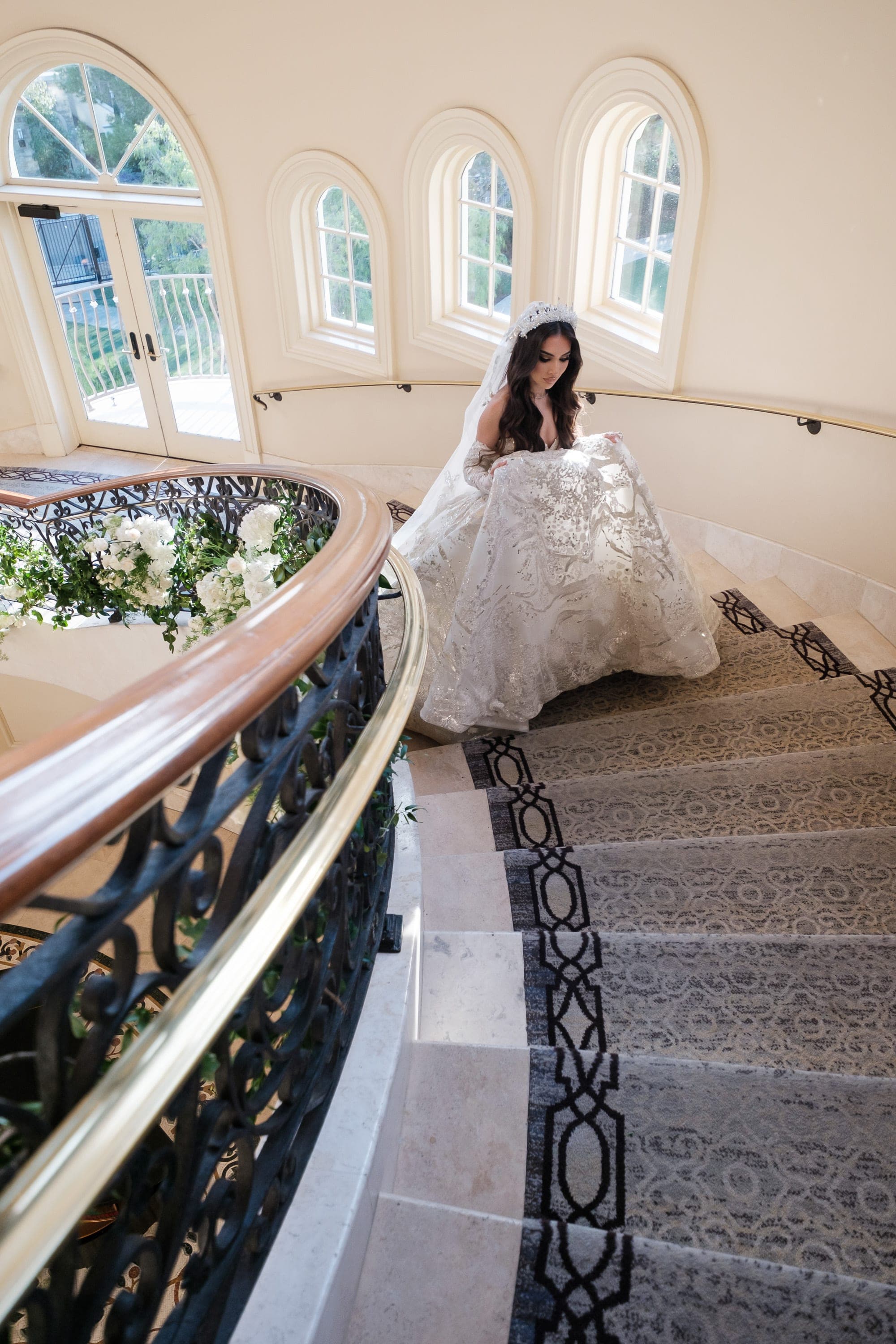 Bride in beaded gown descends a curved staircase, lifting her skirt over patterned carpet.
