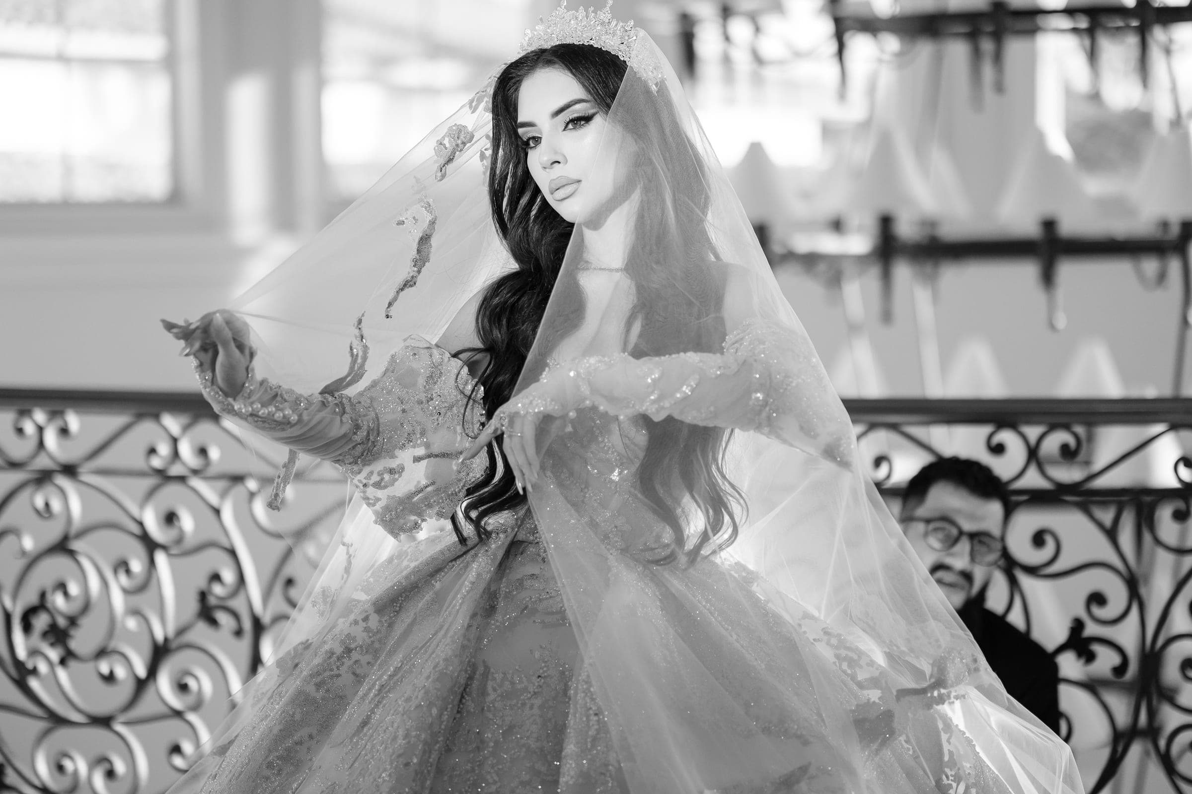 Black and white portrait of bride adjusting her veil while a man watches through iron railing.