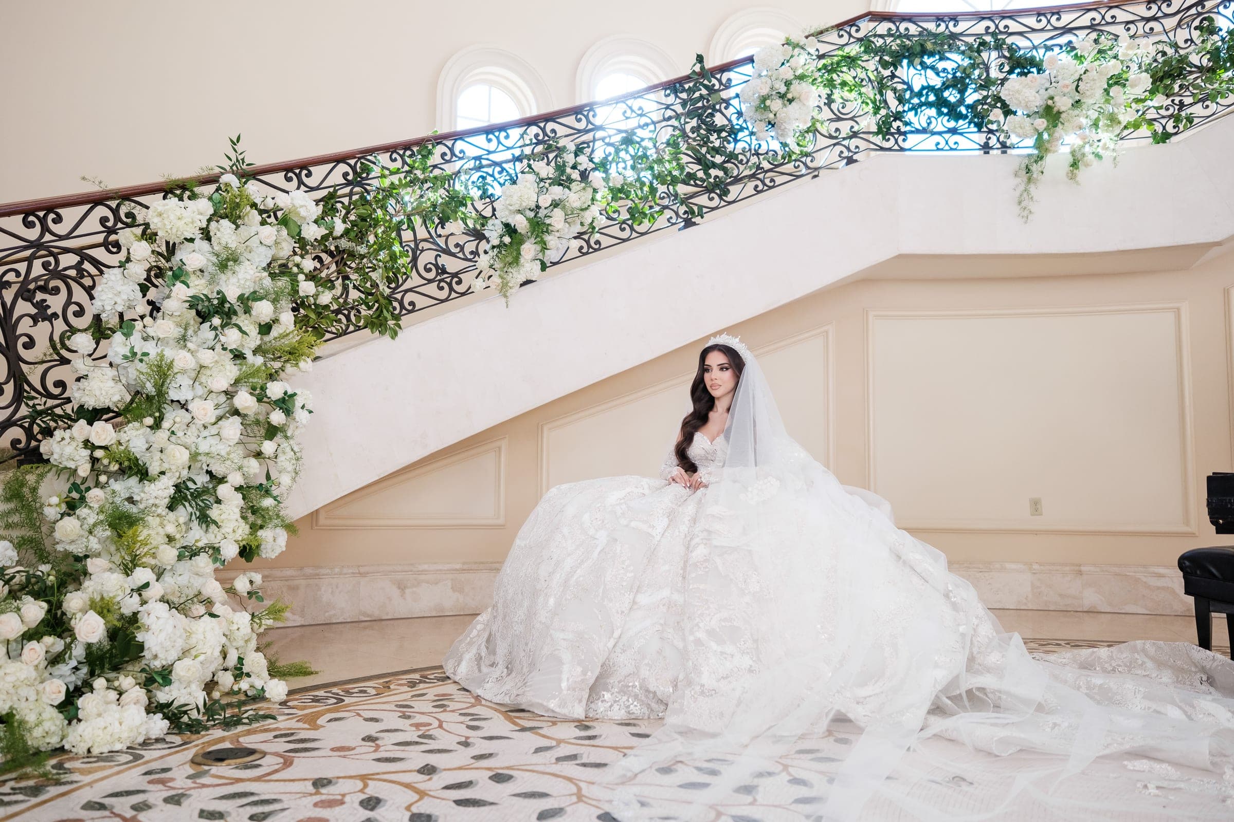 Bride sits at the base of a floral-draped staircase in a voluminous lace ballgown.