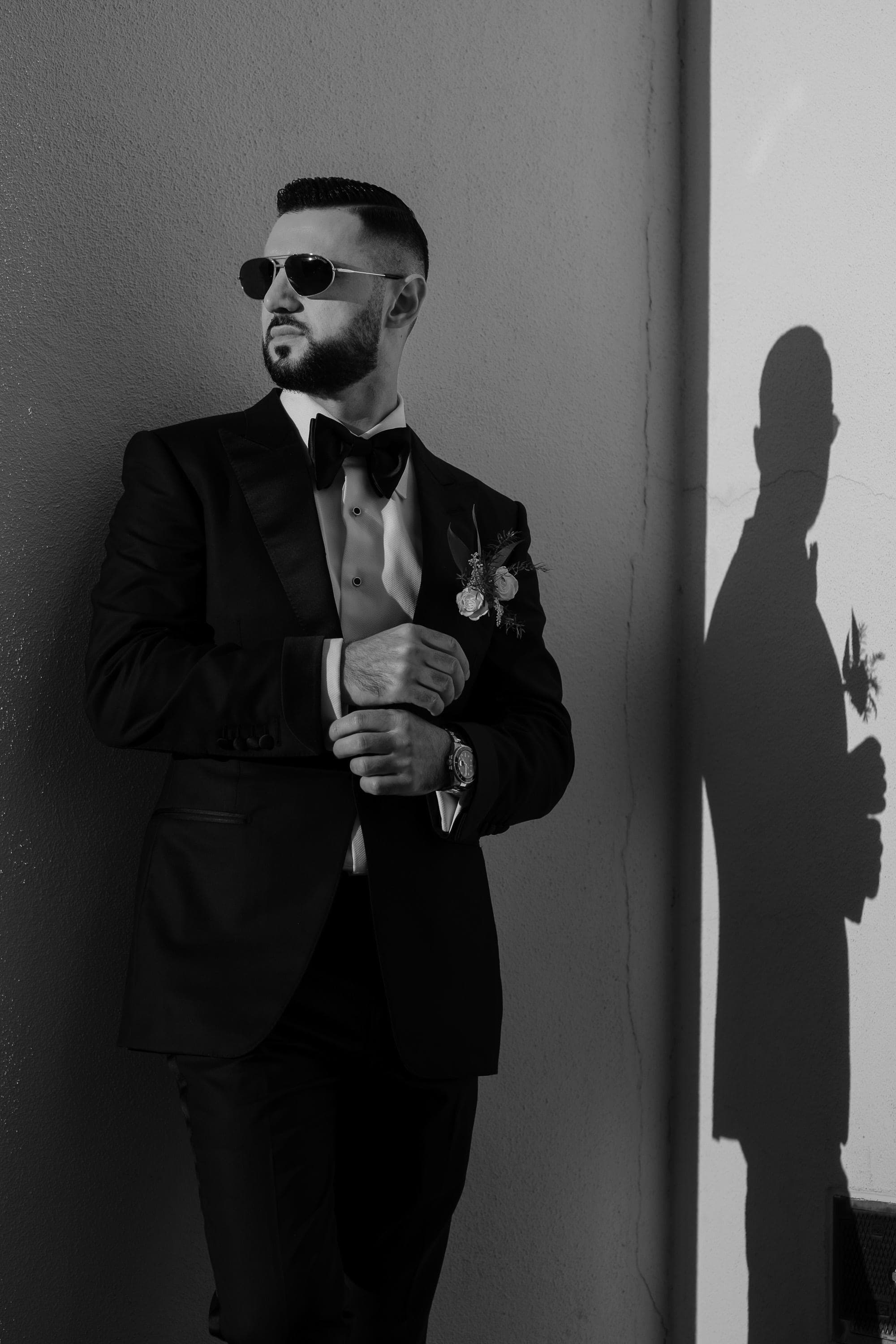 Black and white portrait of a bearded groom in sunglasses and tuxedo leaning against a wall with his shadow beside him.