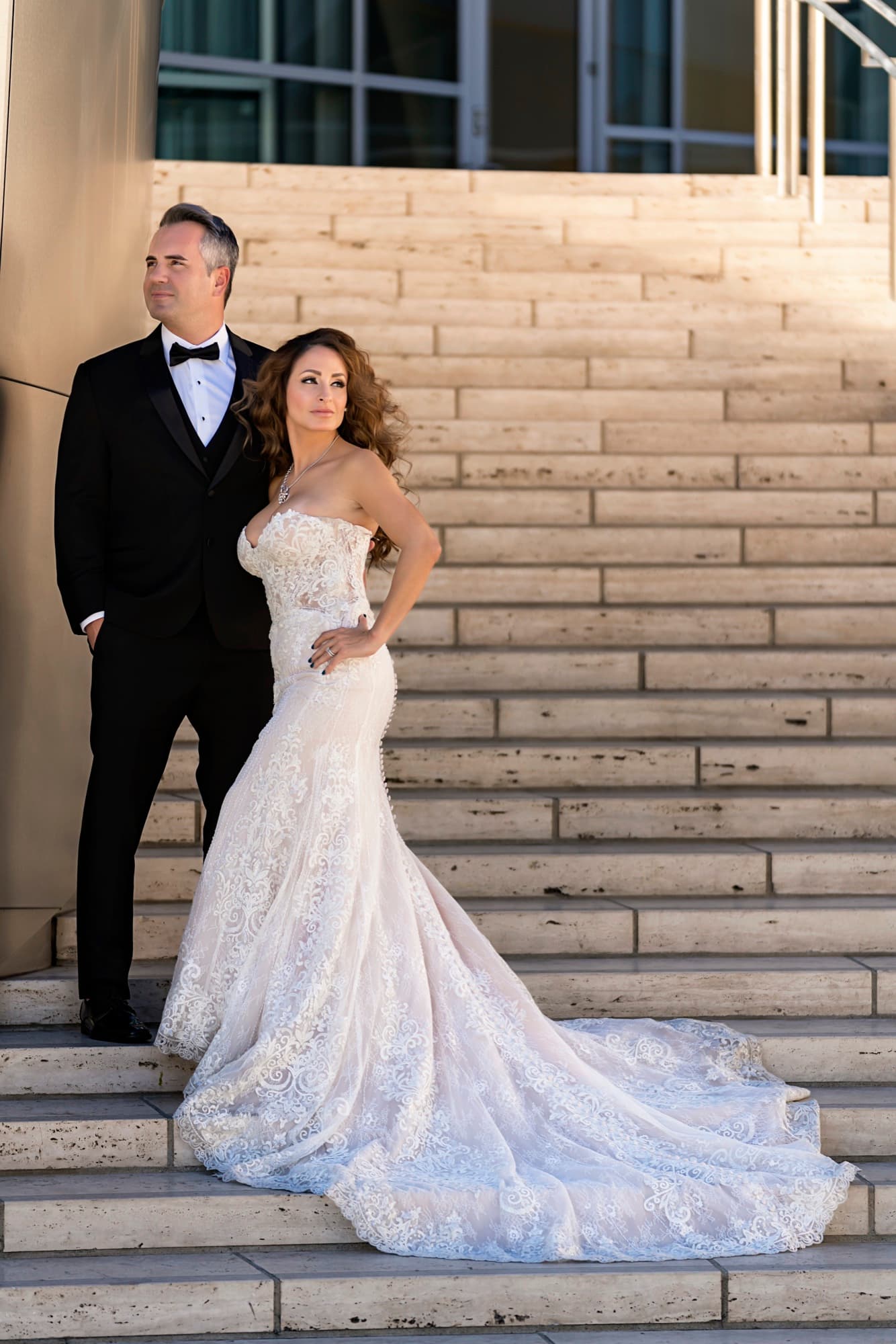 Suzie seated on the travertine grand staircase at Walt Disney Concert Hall in a beaded mermaid gown, Paul standing behind her
