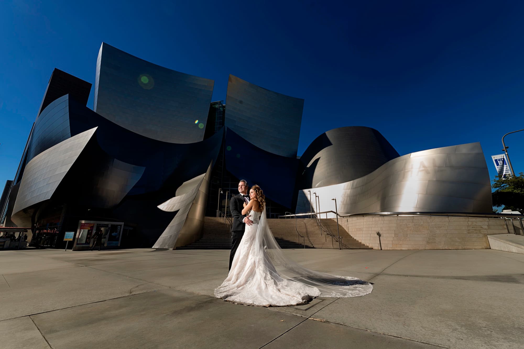 Suzie and Paul embracing in front of the curving steel sails of Walt Disney Concert Hall, her cathedral train sweeping across the plaza, photographed by Michael Anthony Photography