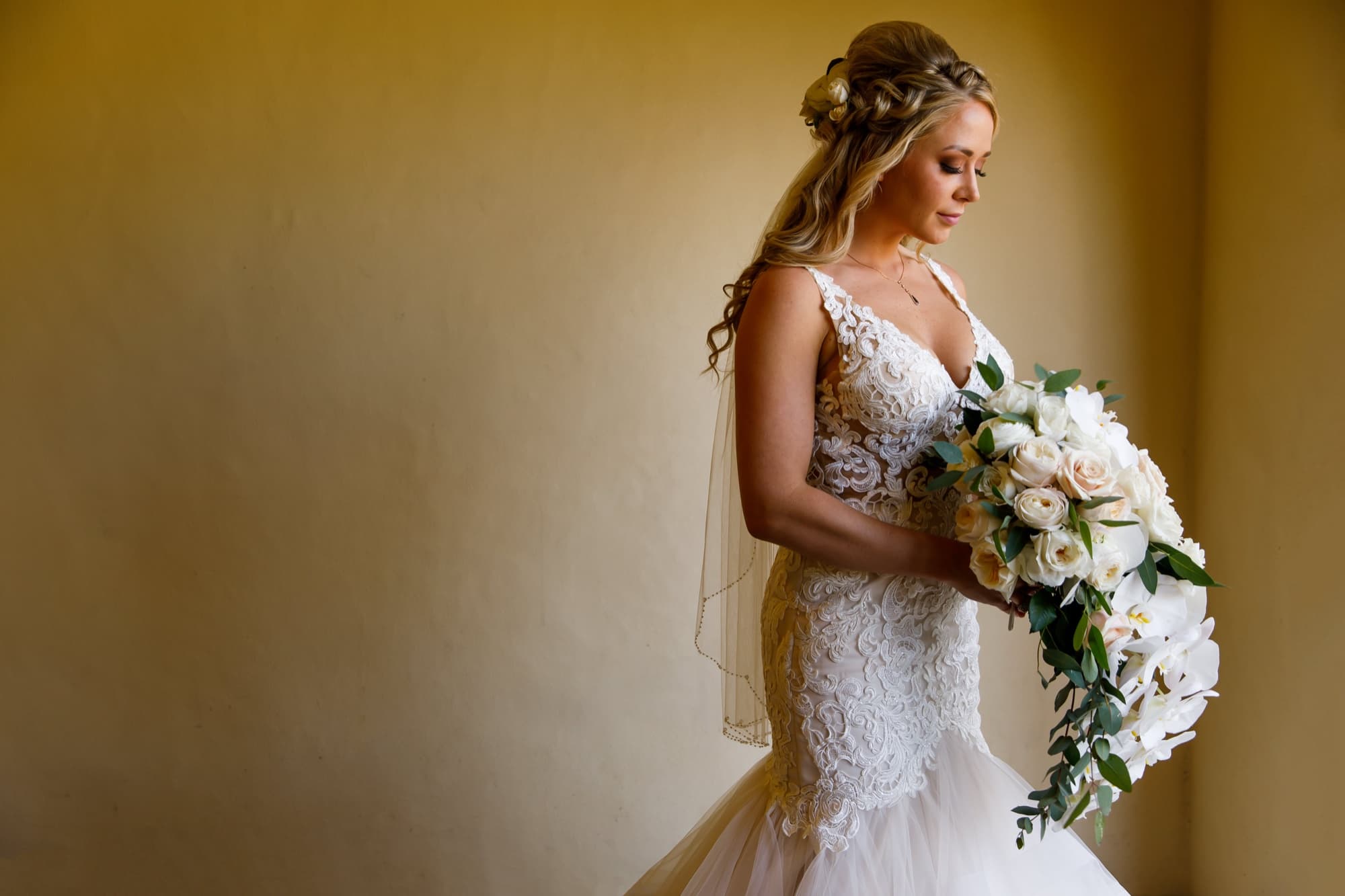 Amanda in a lace mermaid gown holding a cascading white orchid bouquet in the window light before her Wayfarers Chapel wedding