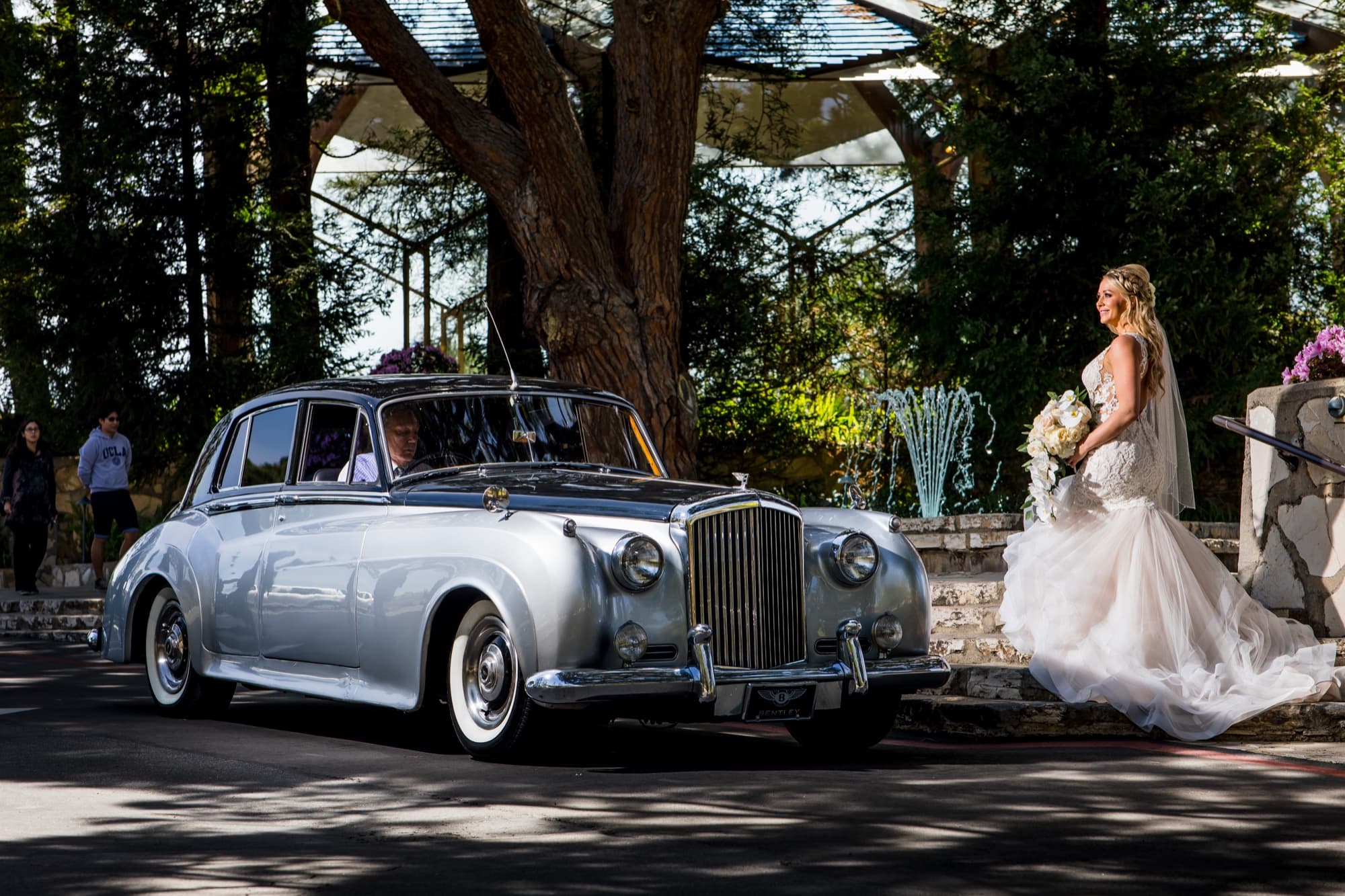Amanda walking past a classic silver saloon car beneath the trees outside Wayfarers Chapel