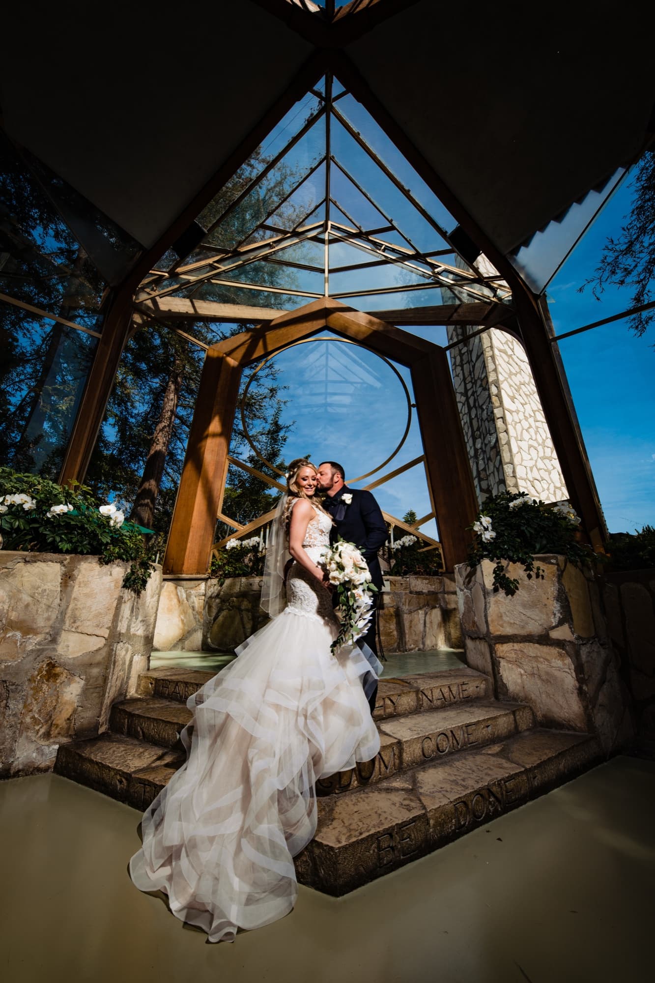 Amanda and Sean kissing on the stone steps inside the glass Wayfarers Chapel in Rancho Palos Verdes, her train cascading down the steps, photographed by Michael Anthony Photography