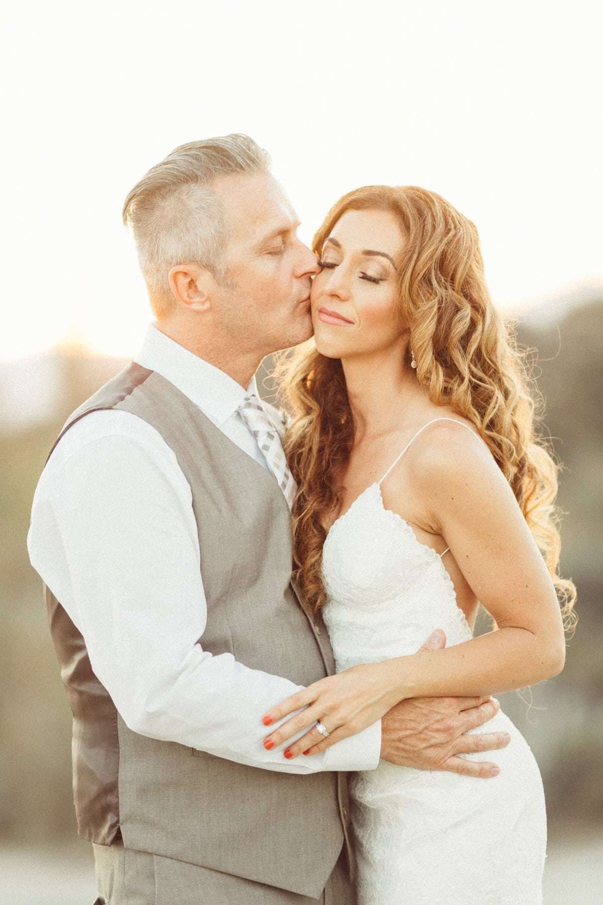 A groom kissing his bride on the forehead in backlit golden light, a compressed telephoto portrait shot on the Canon 135mm f/2L
