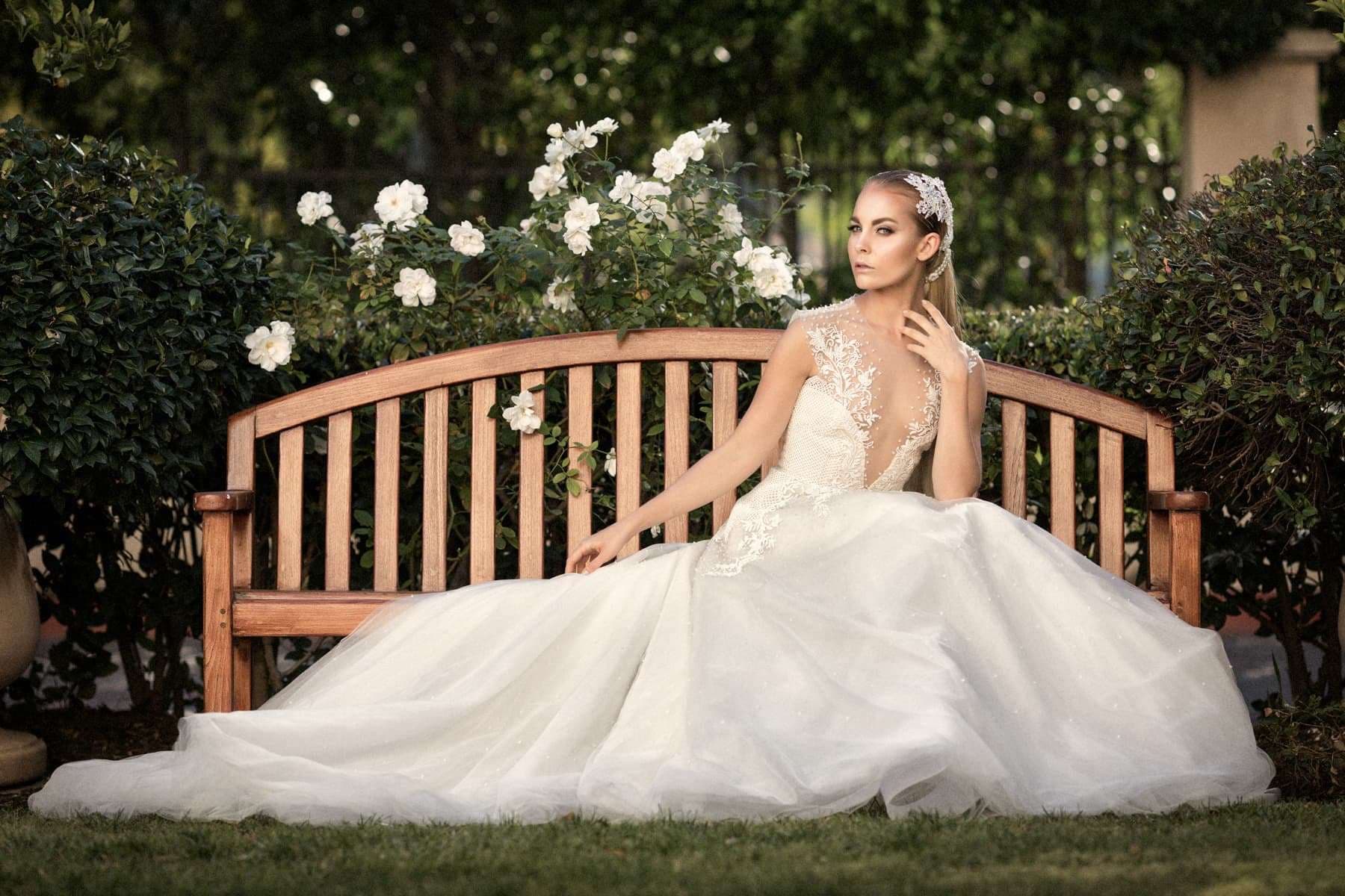 A bride in a full ballgown seated on a wooden garden bench beside white roses, a portrait shot on the Canon 135mm f/2L