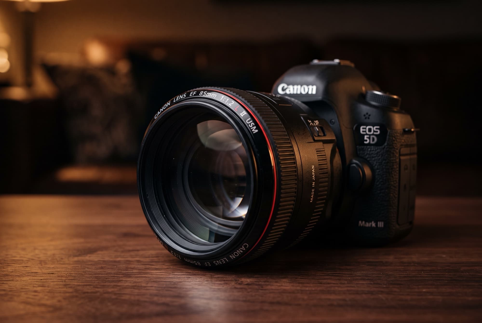 The Canon EF 85mm f/1.2L II USM prime lens mounted on a Canon EOS 5D Mark III body, photographed in moody low light on a dark wood table