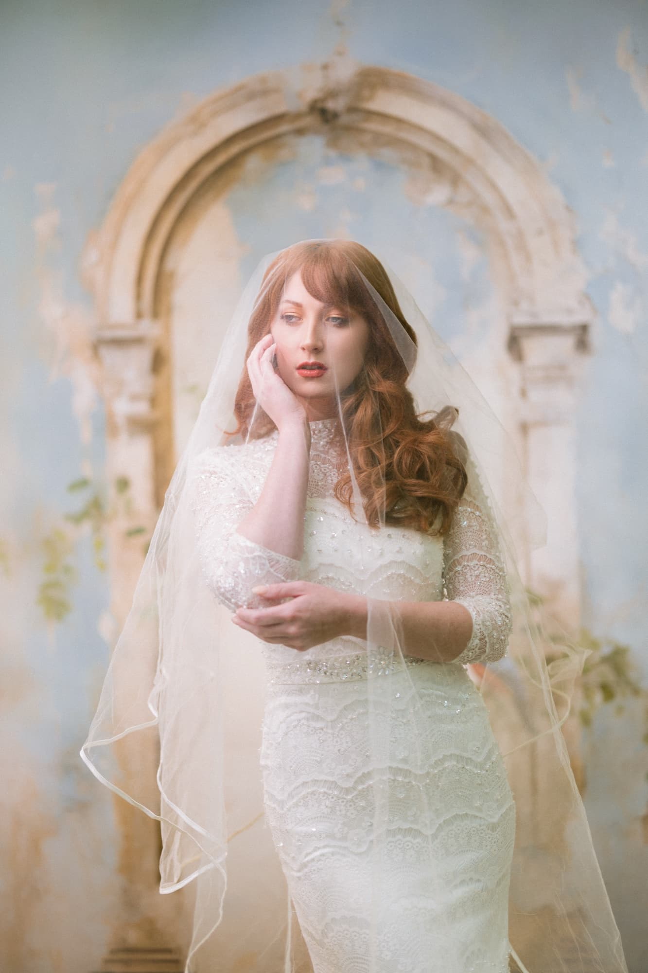 Bride with auburn hair beneath a sheer veil, hand raised to her cheek, photographed tack sharp through the tulle in front of a painterly arch backdrop