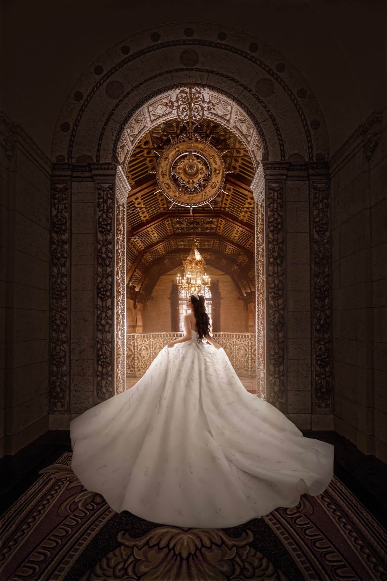 Bride in a cathedral-train ballgown seen from behind, her train fanned across the patterned carpet beneath the ornate arches and chandelier of the Millennium Biltmore
