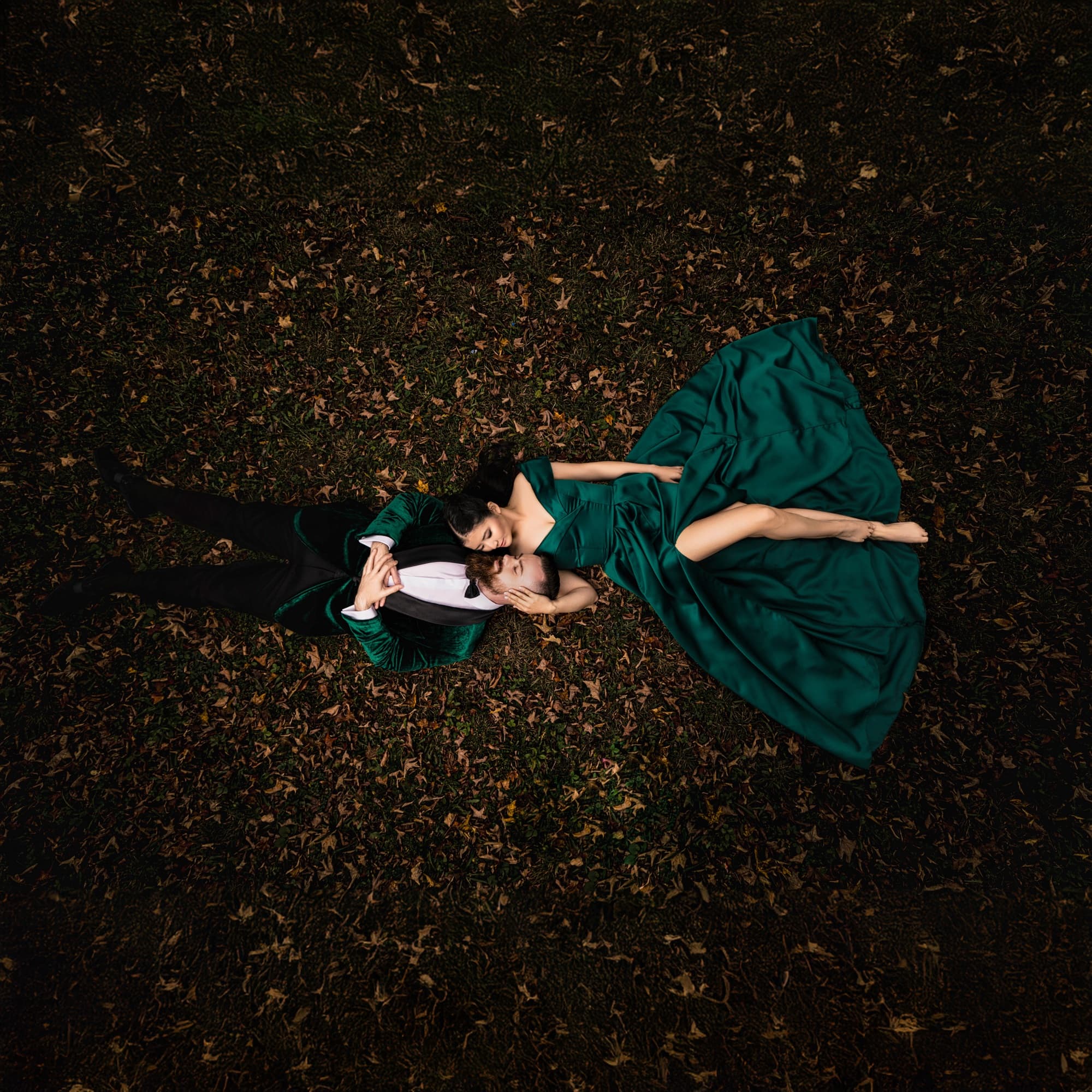 Overhead photograph of a couple lying together in autumn leaves in New York, the woman's emerald satin gown fanned across the dark grass