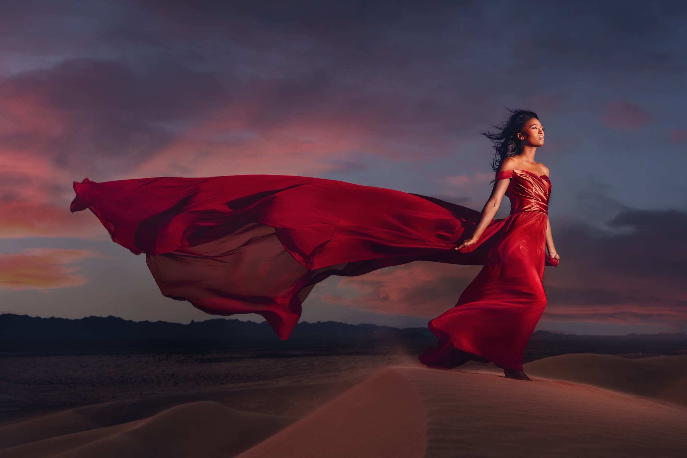 A woman in a red gown on desert dunes at dusk, her dress catching the wind against a stormy pink sky
