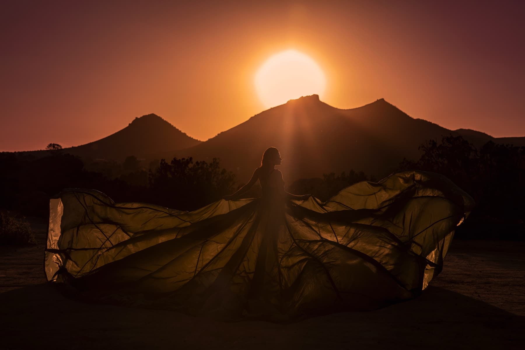 Silhouette of a woman with a sheer train catching the wind in front of a mountain ridge as the sun sets behind it