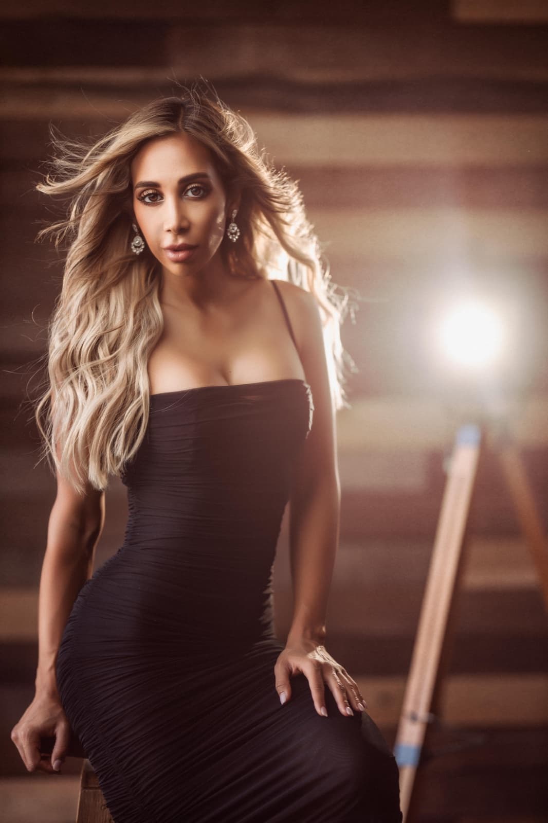 A woman in a black dress lit by a Profoto flash visible on a stand behind her, flaring into the lens in a rustic room