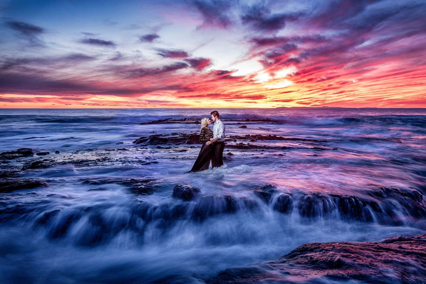 A couple embracing on ocean rocks at sunset with a vivid pink and purple sky over the Pacific in Orange County