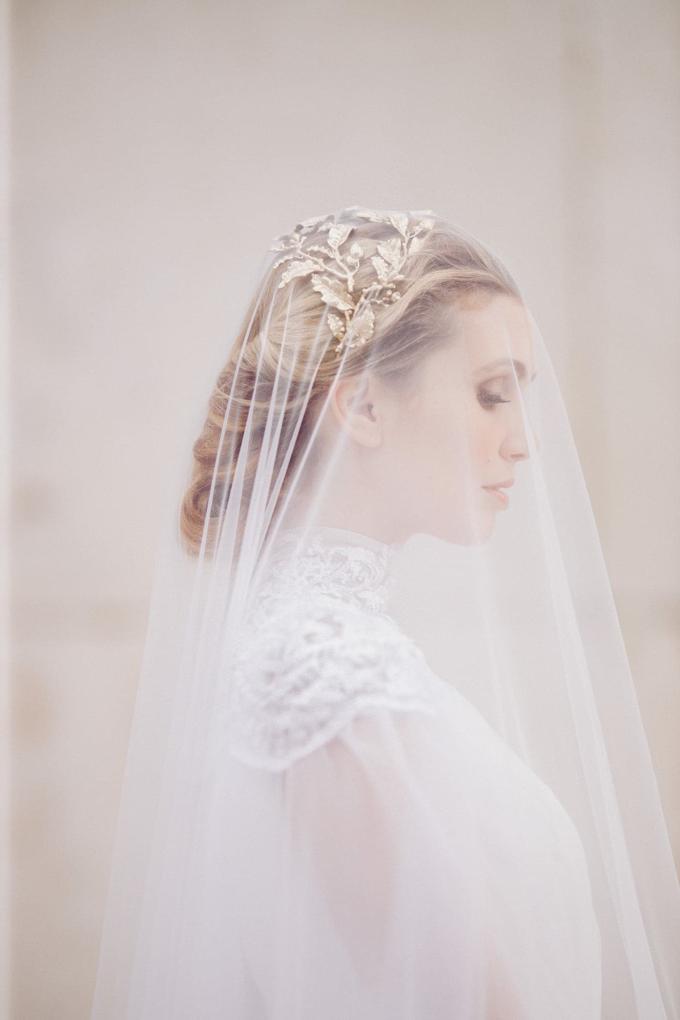 Bride in profile beneath a sheer veil with a gold laurel headpiece, against pale Roman stone